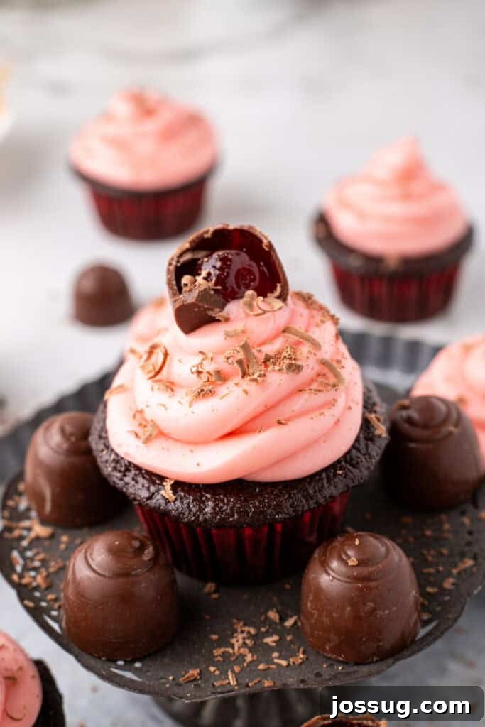 Close up view of a chocolate cupcake topped with pink frosting, chocolate shavings, and a halved cherry cordial candy, surrounded by whole cherry cordial candies