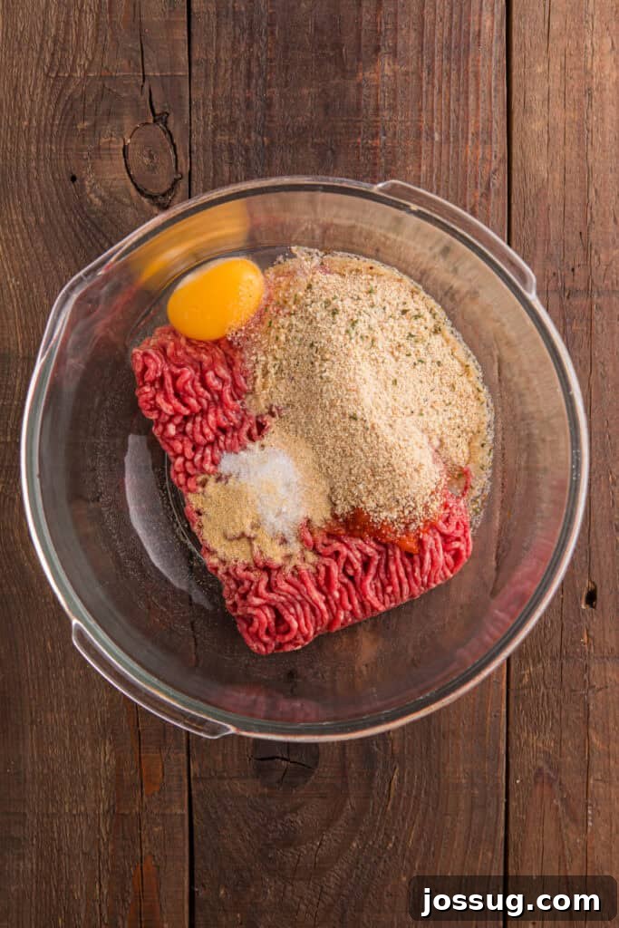 Ground beef, egg, breadcrumbs, chopped onion, ketchup, and seasonings in a clear glass mixing bowl on a wooden surface.