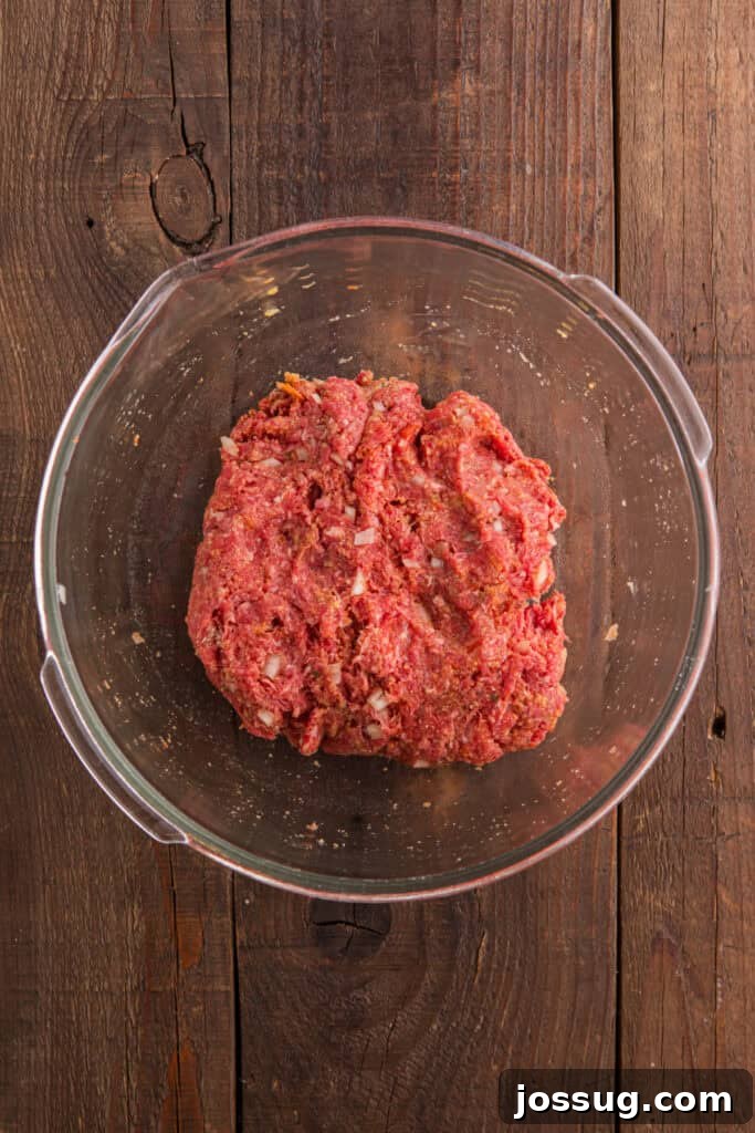 Mixed raw meatloaf mixture with visible chopped onion in a clear glass bowl on a wooden table.