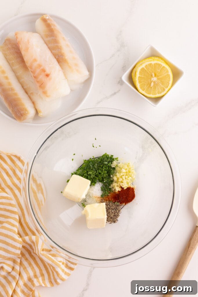 Glass mixing bowl with butter, minced garlic, chopped parsley, paprika, salt, and black pepper, with raw cod fillets and a halved lemon nearby.