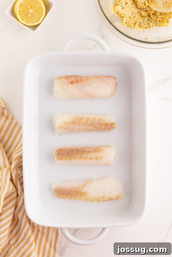 Raw cod fillets arranged in a white baking dish on a marble countertop.