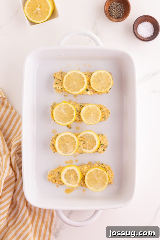 Cod fillets topped with lemon slices and garlic herb butter mixture in a white baking dish before baking.