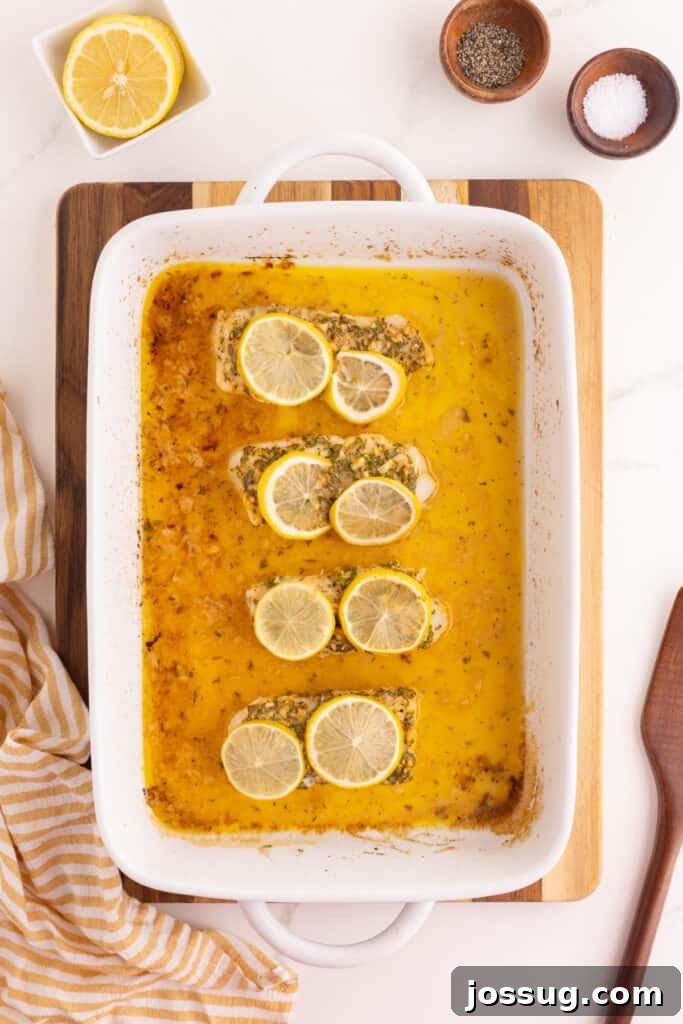 Baked cod fillets in a white baking dish surrounded by melted butter sauce with lemon slices on top.