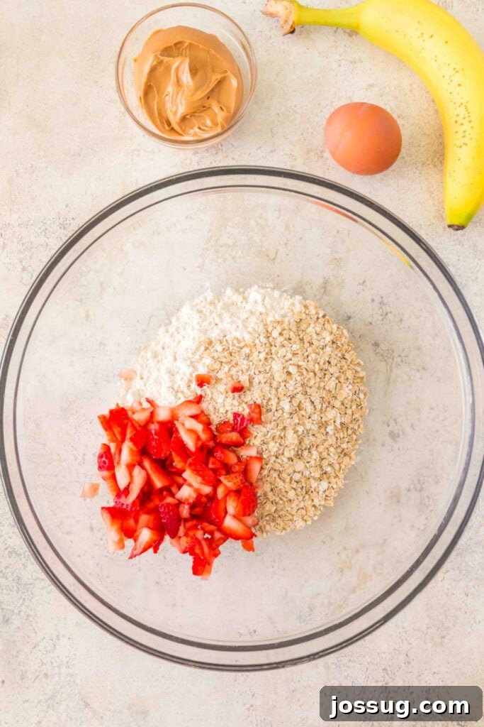 flour, oats, and chopped strawberries in a glass mixing bowl with banana, peanut butter, and egg nearby