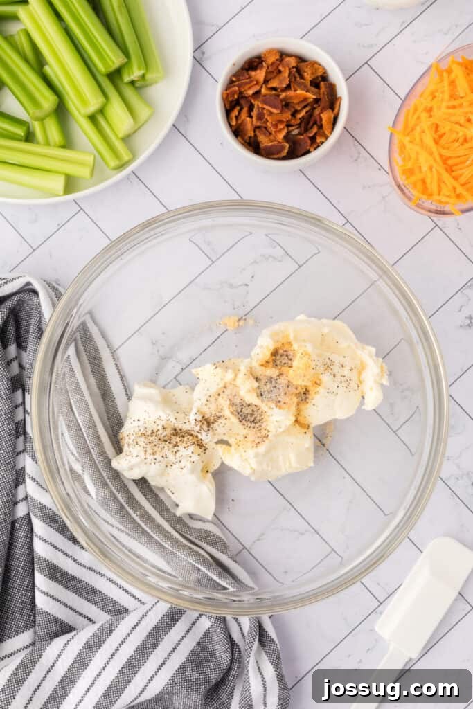 Cream cheese and sour cream with seasonings in a glass mixing bowl next to celery, bacon, and shredded cheddar