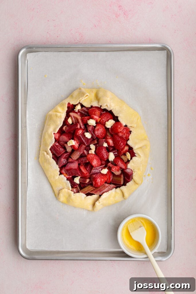 Simply Irresistible Strawberry Rhubarb Galette 17 The unbaked galette crust is being carefully brushed with an egg wash using a pastry brush, ensuring a beautiful golden-brown and shiny finish after baking.