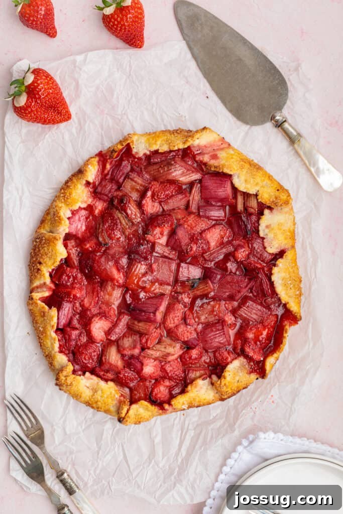 Simply Irresistible Strawberry Rhubarb Galette 20 The exquisite baked strawberry rhubarb galette sits on parchment paper on a wire rack, surrounded by fresh strawberries and baking utensils, cooling and ready to be enjoyed.