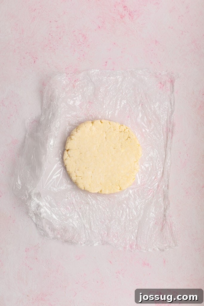 Simply Irresistible Strawberry Rhubarb Galette 9 A neatly flattened disk of galette dough, tightly wrapped in plastic wrap, is chilling on a soft pink surface, an essential step before it can be rolled out.