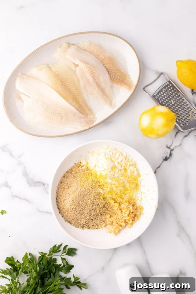 Raw flounder fillets on a plate next to a bowl with breadcrumbs, parmesan, garlic, and lemon zest with lemons and a grater nearby, showing the coating ingredients.