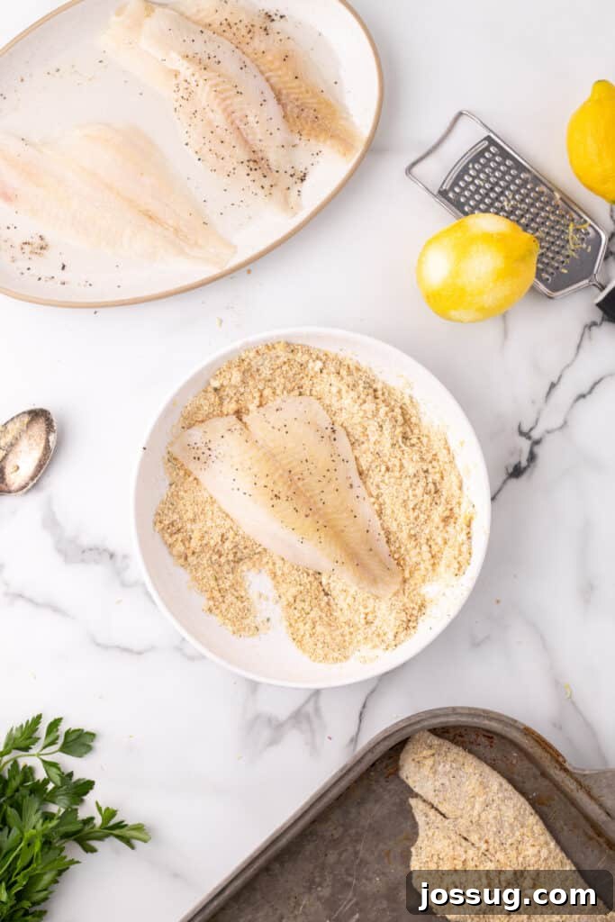 Flounder fillet being coated in breadcrumb and parmesan mixture in a bowl with lemons and grater nearby, demonstrating the coating process.