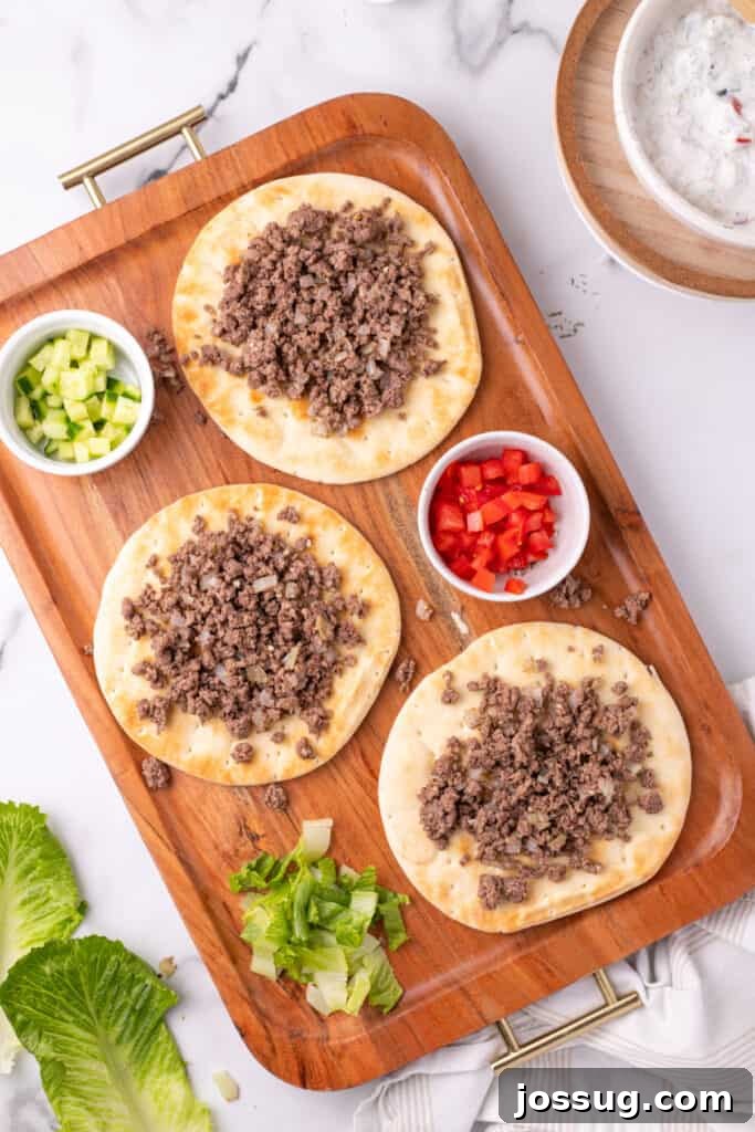 Hafta İçi Akşamlarına Özel Kolay Yunan Kıymalı Pide 11 Pita breads generously filled with seasoned ground beef and topped with creamy yogurt sauce, displayed on a serving tray with an assortment of fresh chopped vegetables.