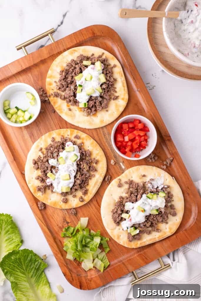 Hafta İçi Akşamlarına Özel Kolay Yunan Kıymalı Pide 12 A close-up of a single pita bread, beautifully topped with ground beef, creamy yogurt sauce, crisp cucumber, juicy tomato, and fresh lettuce, presented on a wooden tray.