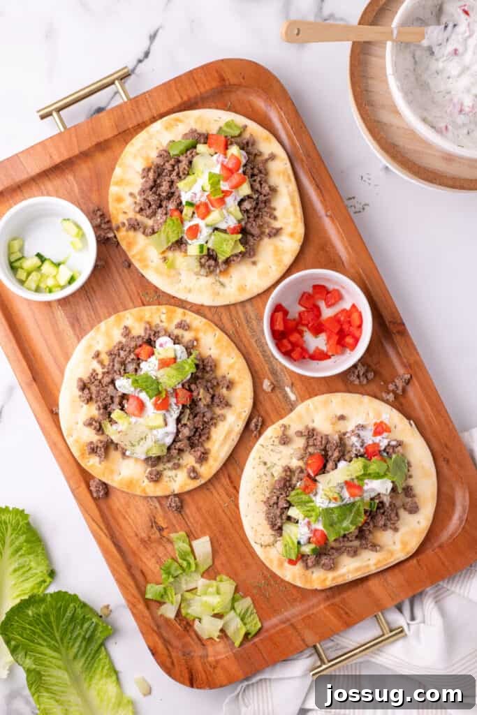 Hafta İçi Akşamlarına Özel Kolay Yunan Kıymalı Pide 14 A detailed close-up of a pita bread topped with ground beef, yogurt sauce, cucumber, tomato, and lettuce, with fries blurred in the background, on a wooden tray.