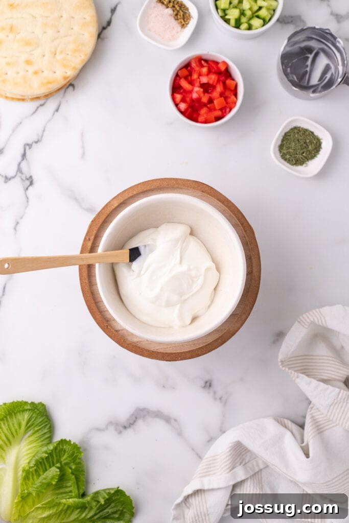 Hafta İçi Akşamlarına Özel Kolay Yunan Kıymalı Pide 6 A bowl of plain Greek yogurt with a spatula, surrounded by fresh chopped vegetables and pita bread on a marble surface, ready for sauce preparation.