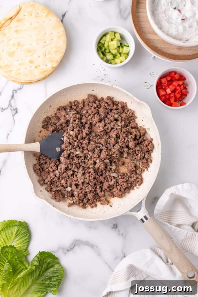 Hafta İçi Akşamlarına Özel Kolay Yunan Kıymalı Pide 9 Cooked ground beef and onions in a skillet with a spatula, fully browned and seasoned, on a marble surface.