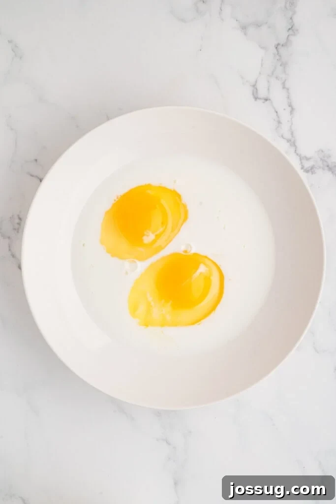 Effortless Crispy Chicken Strips for Busy Families 7 A simple yet essential image of two large eggs cracked into a bowl, alongside a splash of whole milk, poised and ready to be whisked together for the wet dredging mixture.