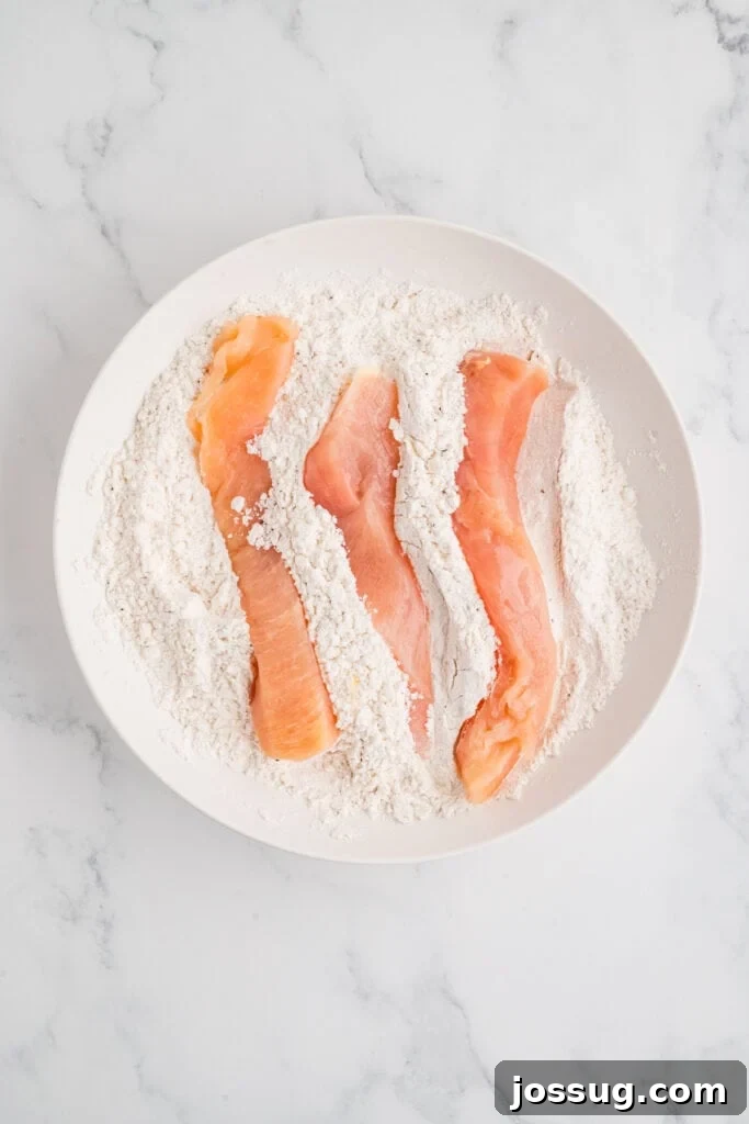 Effortless Crispy Chicken Strips for Busy Families 8 Chicken strips uniformly coated in the first layer of seasoned flour, carefully arranged and resting in a bowl, awaiting their dip into the egg mixture.