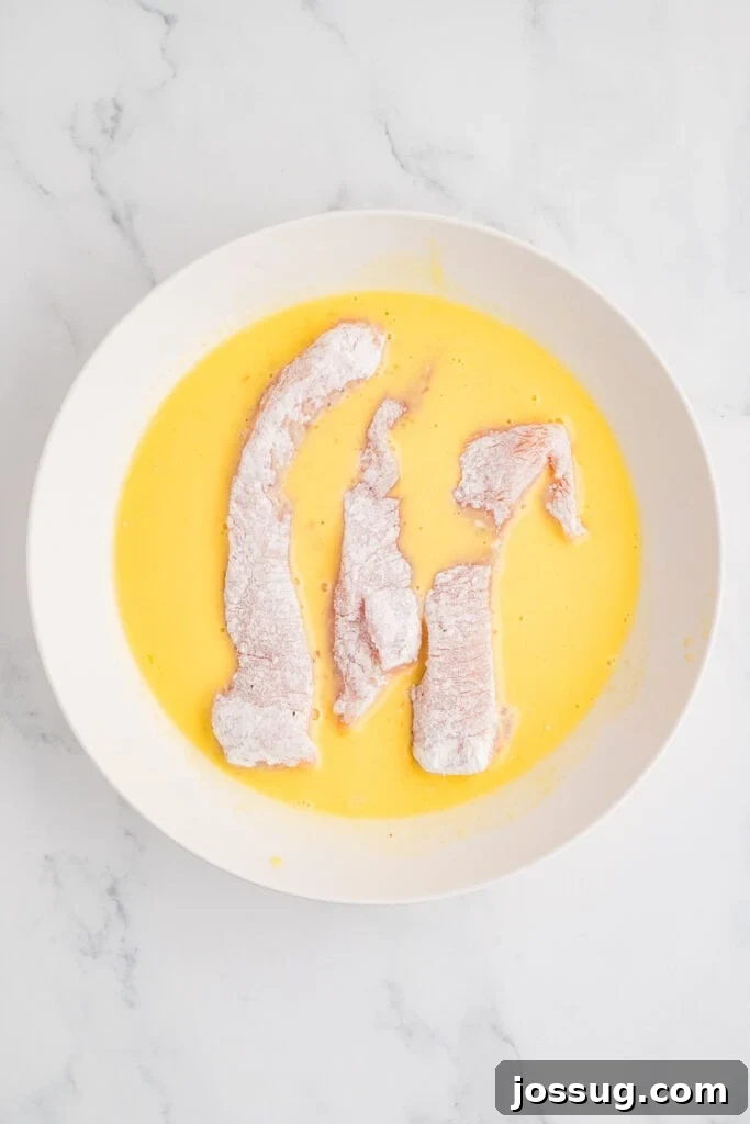 Effortless Crispy Chicken Strips for Busy Families 9 A close-up shot of a floured chicken strip being carefully dipped into the smooth egg and milk mixture, ensuring an even coating before the final layer of flour.