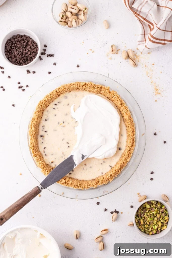 Dreamy No Bake Cannoli Pie with Ricotta and Chocolate Chips 11 Overhead view of whipped cream being spread onto a no-bake cannoli pie in a glass pie dish, with bowls of mini chocolate chips and chopped pistachios nearby.