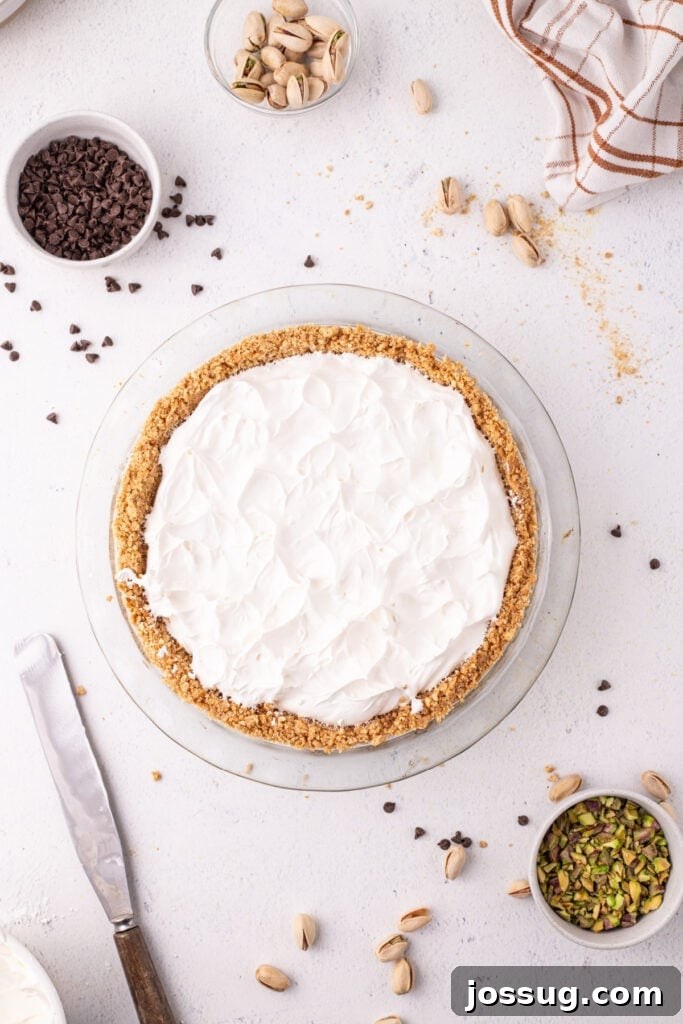 Dreamy No Bake Cannoli Pie with Ricotta and Chocolate Chips 12 Overhead view of a no-bake cannoli pie with whipped cream fully spread across the top in a glass pie dish, surrounded by mini chocolate chips, pistachios, and a linen towel.