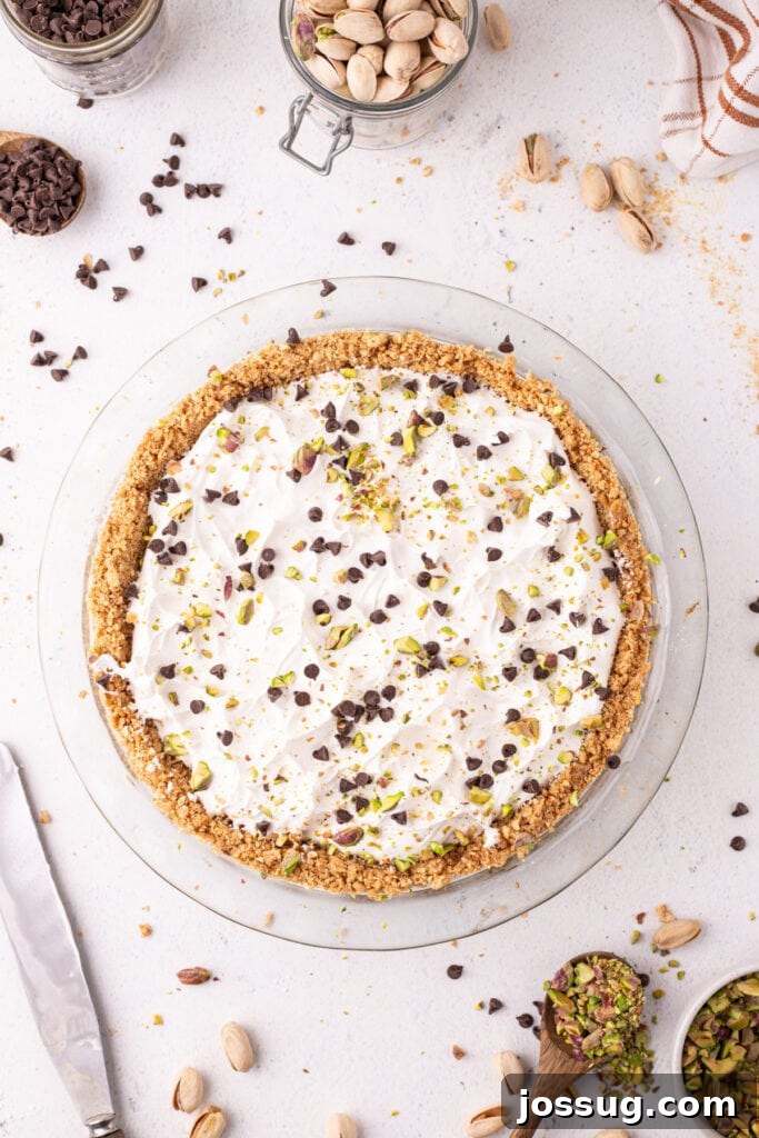 Overhead view of a finished no-bake cannoli pie topped with whipped cream, mini chocolate chips, and chopped pistachios in a glass pie dish, with a jar of pistachios and bowls of toppings around it.