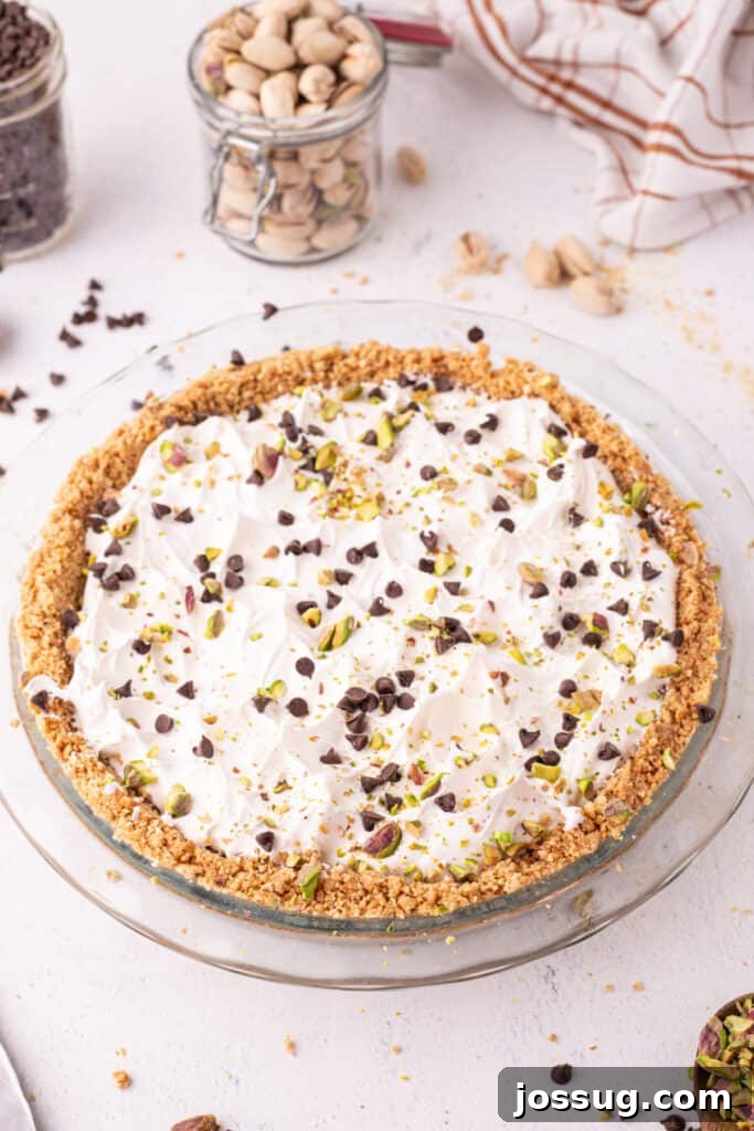 Dreamy No Bake Cannoli Pie with Ricotta and Chocolate Chips 14 Angled view of a finished no-bake cannoli pie in a glass pie dish, topped with whipped cream, mini chocolate chips, and chopped pistachios, with a jar of pistachios and a striped linen towel in the background.