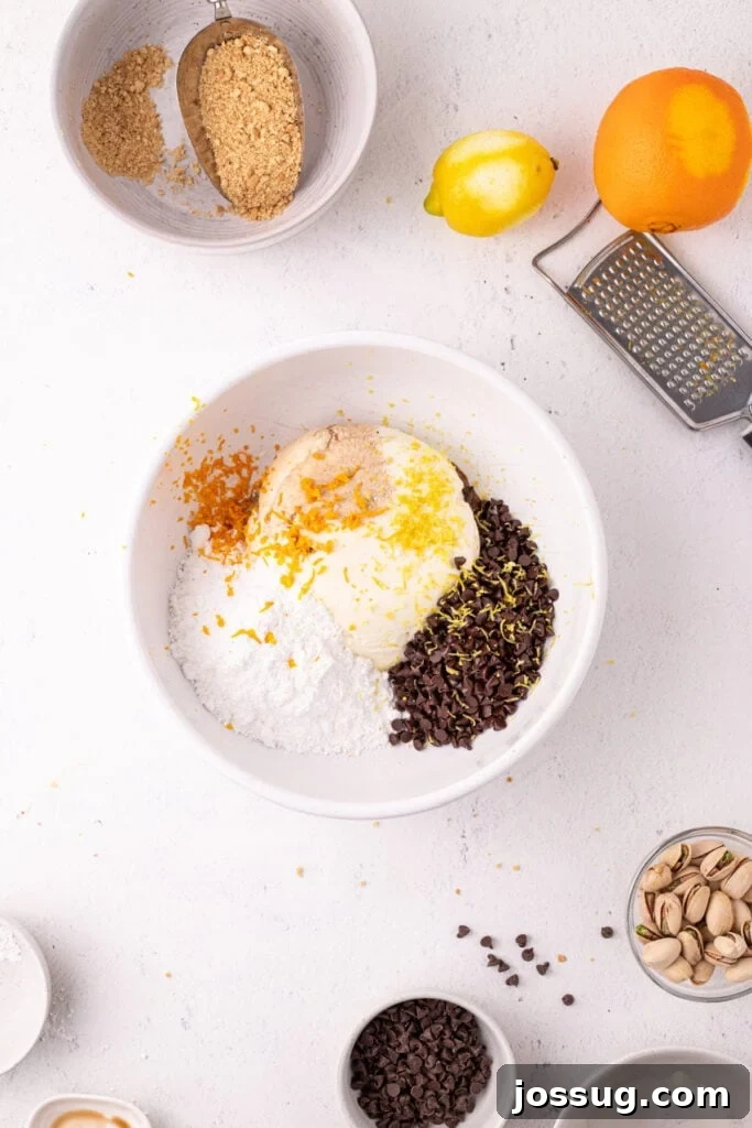 Dreamy No Bake Cannoli Pie with Ricotta and Chocolate Chips 8 Overhead view of a white bowl with ricotta cheese, powdered sugar, mini chocolate chips, and fresh lemon and orange zest, with a box grater and whole citrus fruits nearby.