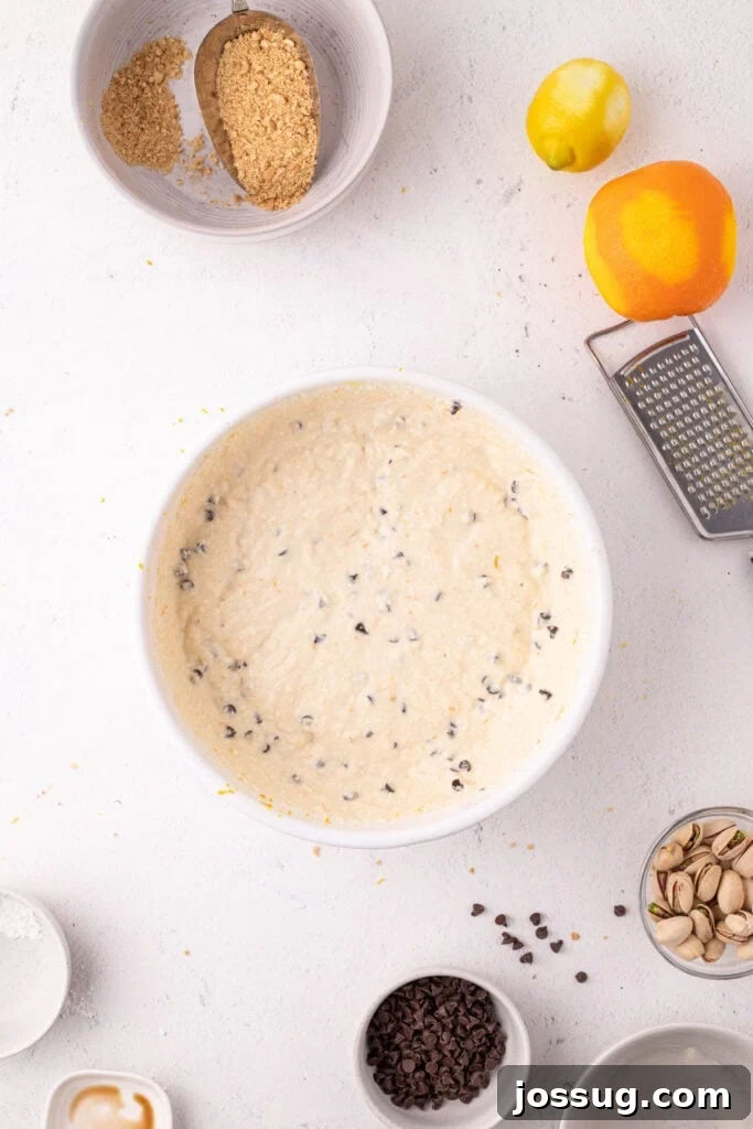 Dreamy No Bake Cannoli Pie with Ricotta and Chocolate Chips 9 Overhead view of the smooth, creamy cannoli filling with mini chocolate chips mixed in, in a white bowl, with a lemon, orange, box grater, pistachios, and chocolate chips arranged around it.