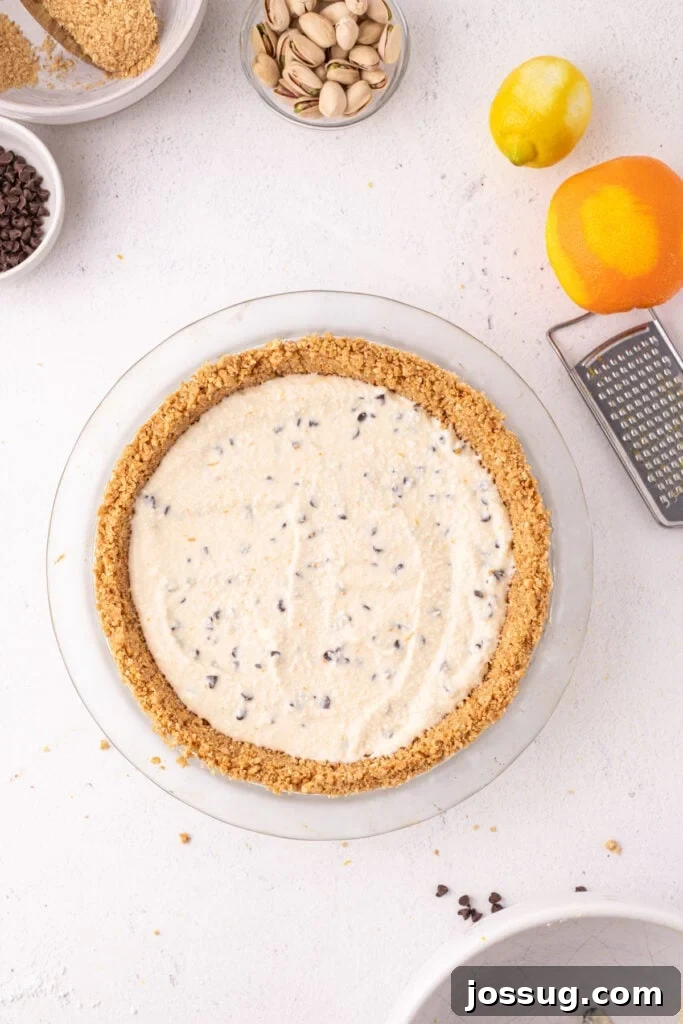 Dreamy No Bake Cannoli Pie with Ricotta and Chocolate Chips 10 Overhead view of creamy cannoli filling spread into a graham cracker crust in a glass pie dish, with a lemon, orange, box grater, pistachios, and mini chocolate chips surrounding it.
