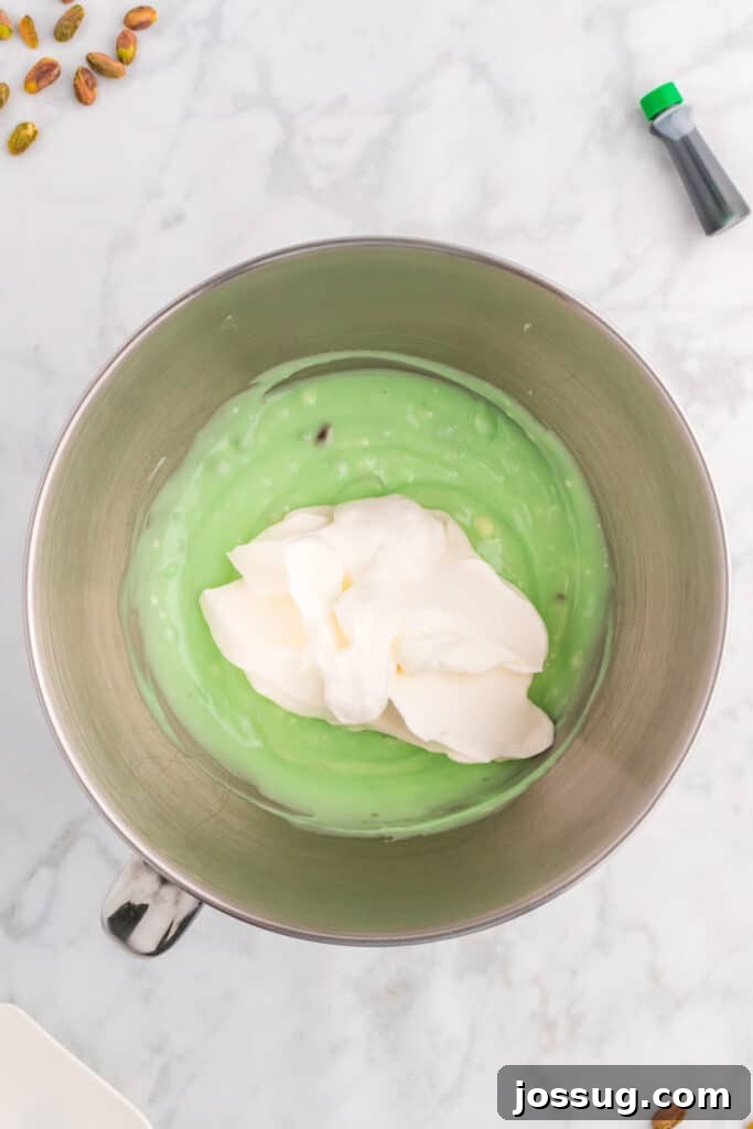 Quick No Bake Pistachio Delight 11 Fluffy whipped cream is carefully added to the pistachio and cream cheese mixture in a mixing bowl, poised for gentle folding to create a light and airy pie filling.