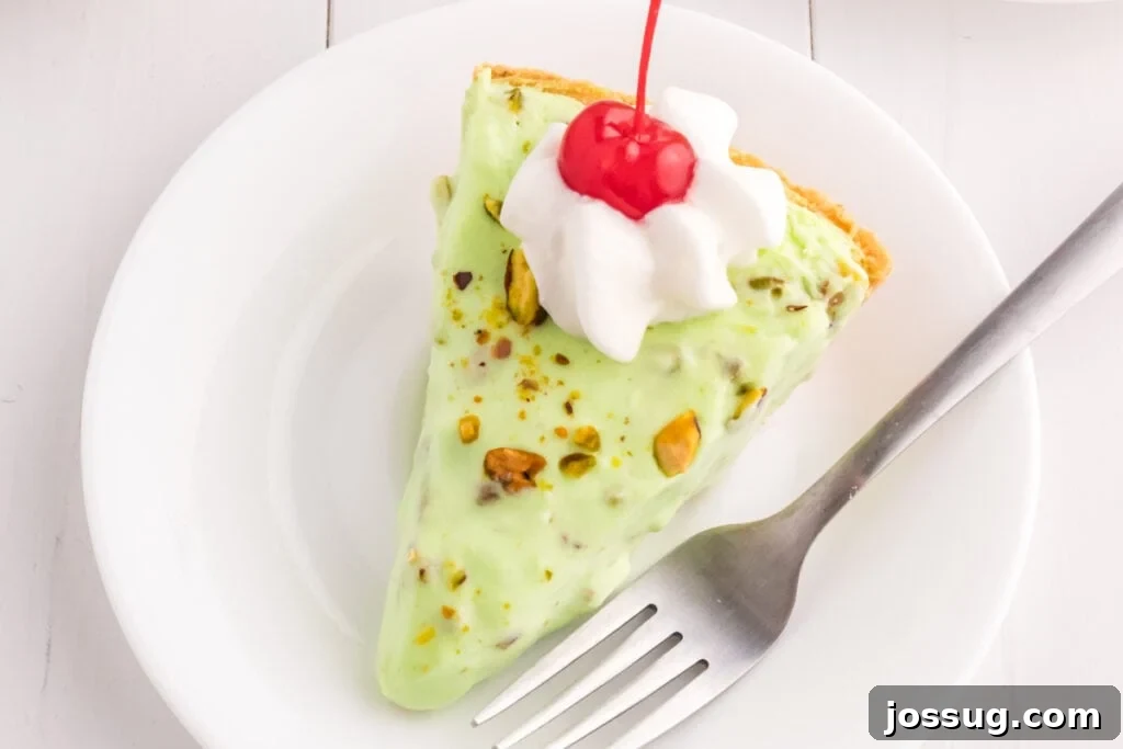 Quick No Bake Pistachio Delight 4 A perfectly portioned slice of No Bake Pistachio Cream Pie, elegantly topped with a dollop of fluffy whipped cream and a bright red maraschino cherry, resting on a pristine white plate.