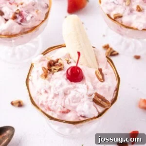 Banana Split Salad served in a glass dessert dish and topped with a banana half, maraschino cherry, coconut, and pecans, with additional dessert dishes in the background.