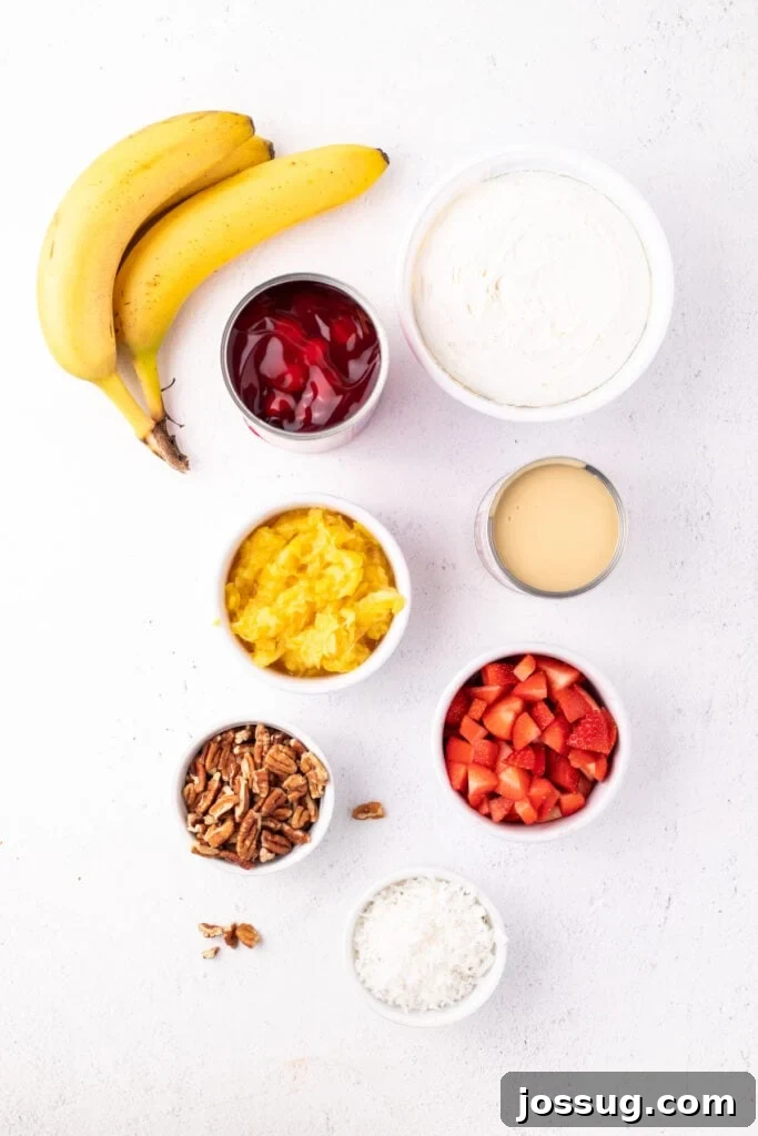 Ingredients for Banana Split Salad arranged on a white surface, including bananas, cherry pie filling, whipped topping, sweetened condensed milk, crushed pineapple, strawberries, pecans, and coconut.