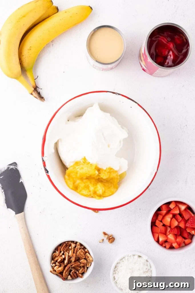 Whipped topping and crushed pineapple in a mixing bowl with bananas, sweetened condensed milk, cherry pie filling, strawberries, pecans, and coconut nearby for Banana Split Salad.