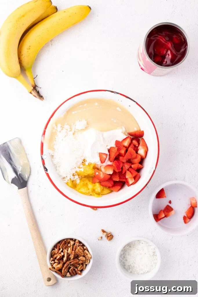 Ingredients for Banana Split Salad in a mixing bowl, including whipped topping, sweetened condensed milk, crushed pineapple, coconut, and chopped strawberries.