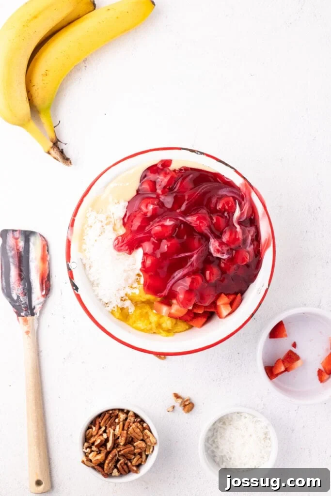 Cherry pie filling added to a mixing bowl with whipped topping, sweetened condensed milk, pineapple, strawberries, and coconut for Banana Split Salad.