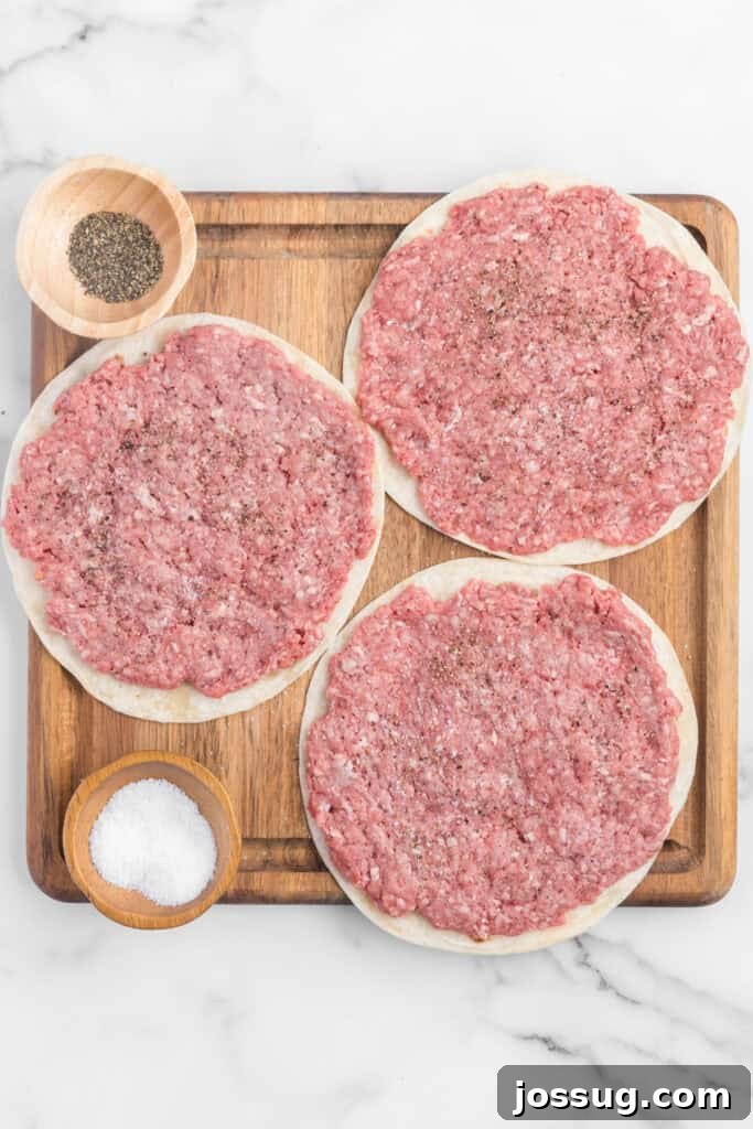 Smash Burger Tacos: Viral Cheeseburger-Style Taco Recipe 8 Flour tortillas topped with a thin layer of raw ground beef and seasoned with salt and pepper on a wooden cutting board.