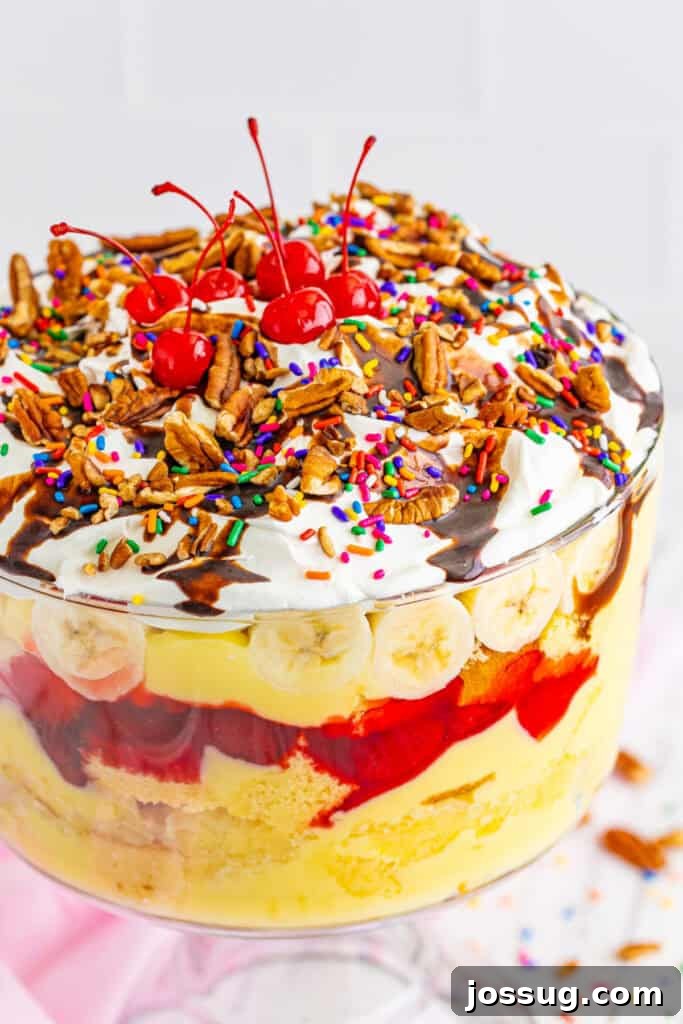 Decadent Banana Split Trifle Recipe with Layers of Cream and Fruit 2 Close-up of finished Banana Split Trifle showing layers of cake, pudding, pineapple, strawberry filling, bananas, whipped topping, chocolate sauce, pecans, sprinkles, and cherries.
