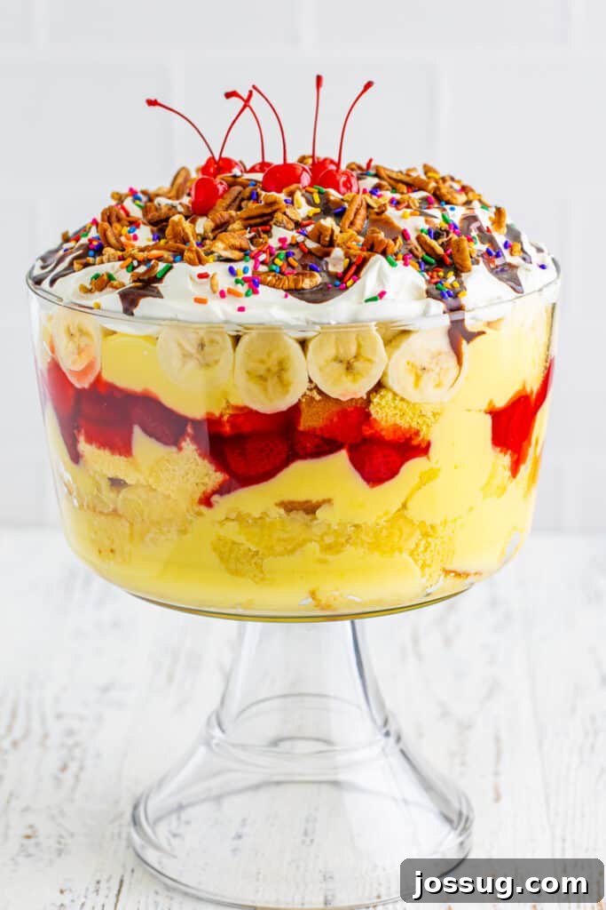 Decadent Banana Split Trifle Recipe with Layers of Cream and Fruit 17 Finished Banana Split Trifle topped with whipped topping, chocolate sauce, chopped pecans, rainbow sprinkles, and maraschino cherries in a clear trifle bowl.