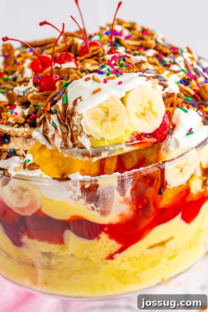 Decadent Banana Split Trifle Recipe with Layers of Cream and Fruit 18 Spoon lifting a serving of Banana Split Trifle from the trifle bowl, showing layers of cake, pudding, bananas, strawberry filling, whipped topping, chocolate sauce, pecans, sprinkles, and cherries.