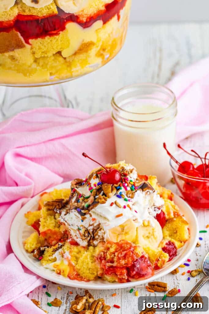 Decadent Banana Split Trifle Recipe with Layers of Cream and Fruit 19 Serving of Banana Split Trifle on a plate topped with whipped topping, chocolate sauce, pecans, rainbow sprinkles, and a maraschino cherry, with the trifle bowl in the background.