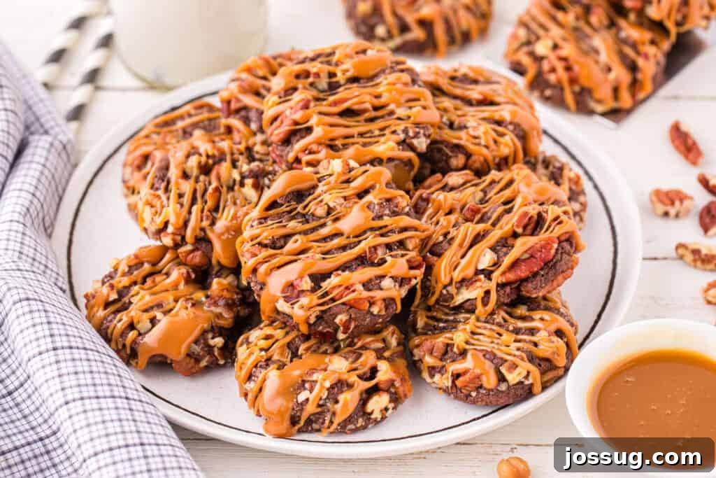 Easy Turtle Cookies piled on a white plate and drizzled with caramel, featuring chocolate cookies coated in chopped pecans and served with a bowl of caramel sauce.