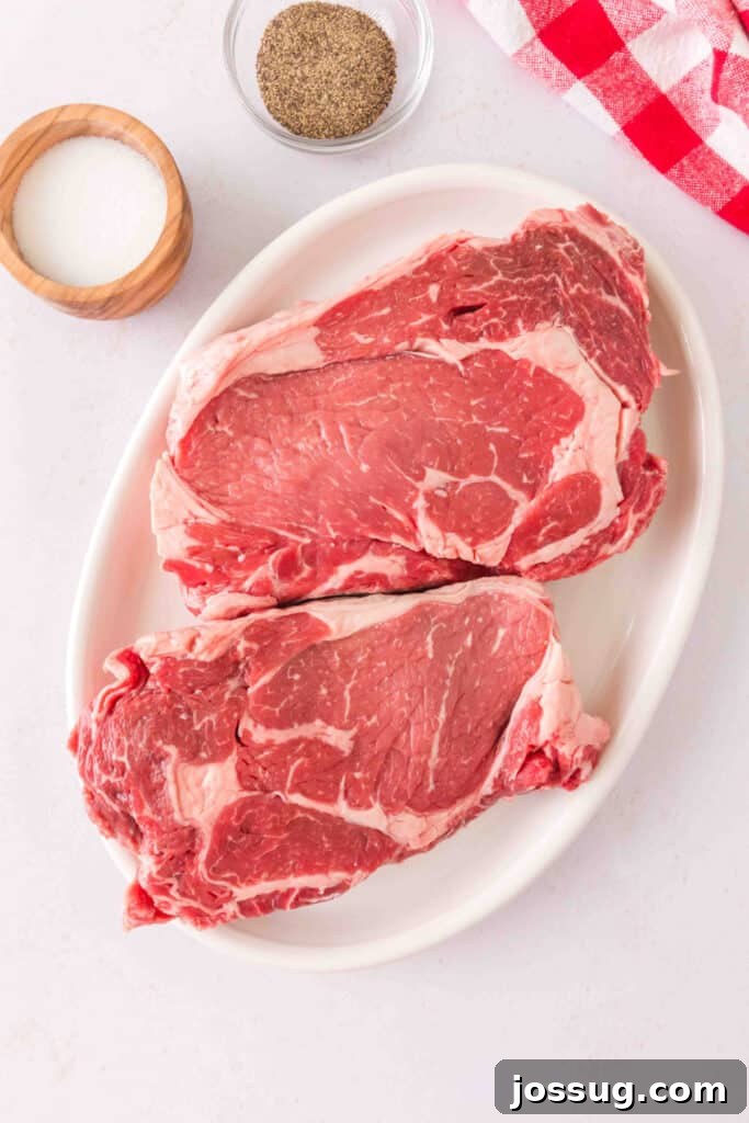 Raw ribeye steaks on a white platter with salt and ground black pepper ready for seasoning.