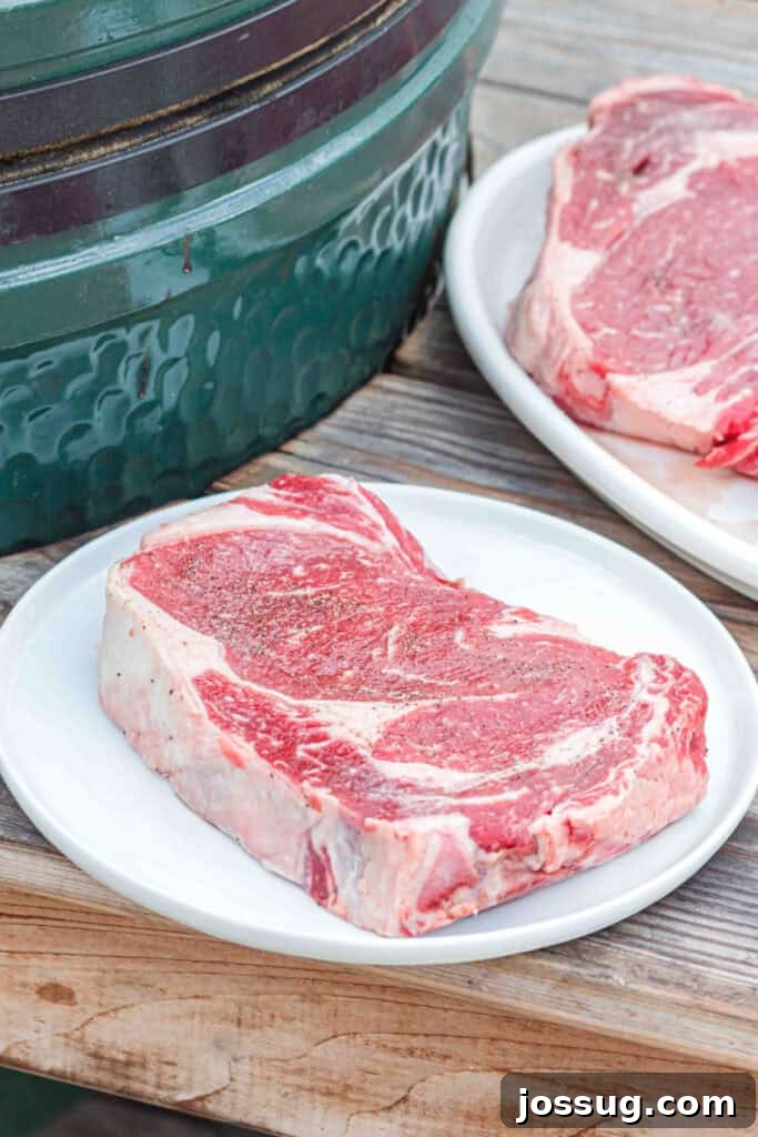 Thick ribeye steak seasoned with salt and pepper beside a BBQ grill.