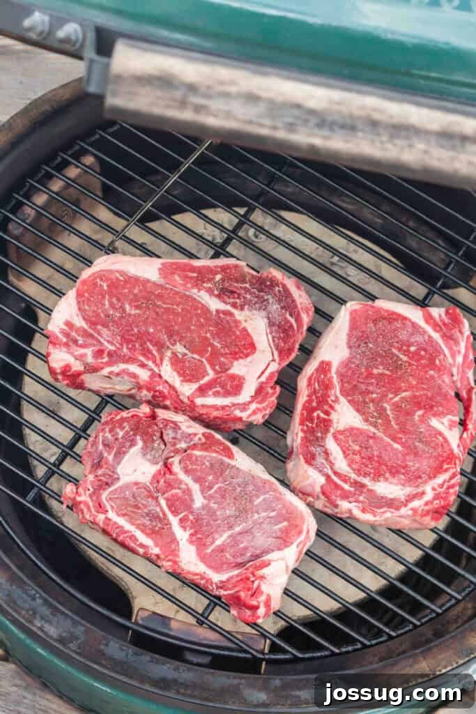 Three seasoned ribeye steaks cooking over indirect heat on a BBQ grill.