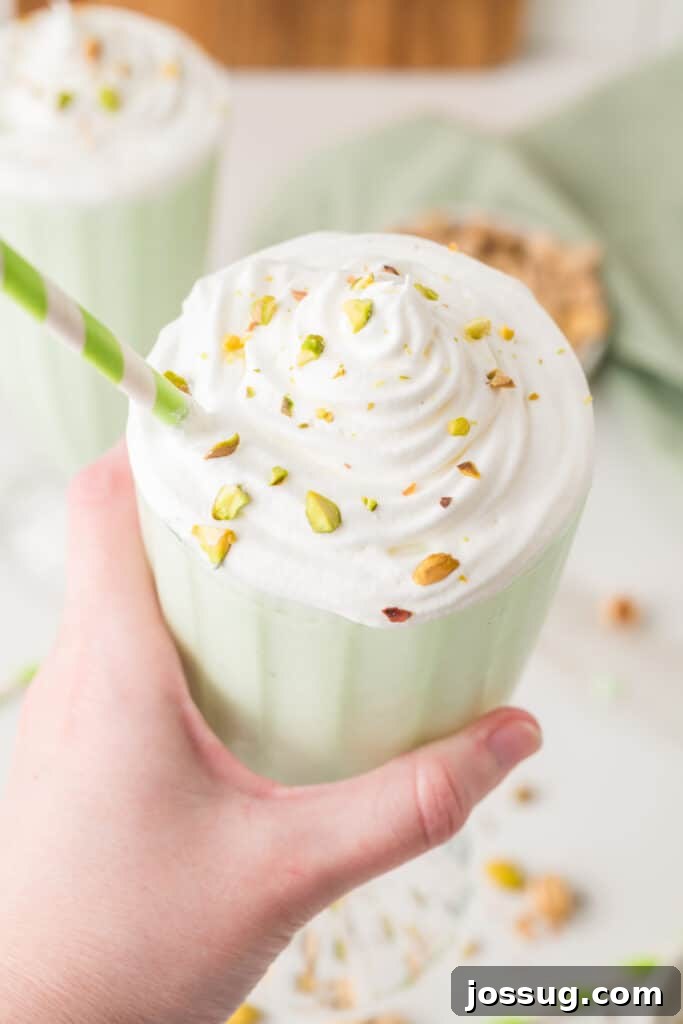 Hand holding a pistachio milkshake topped with whipped cream, chopped pistachios, and a striped straw