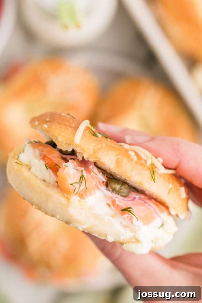 Baked Bagel and Smoked Salmon Sliders 20 Hand holding a Baked Bagels & Lox Slider filled with smoked salmon, cream cheese, red onion, capers, and dill
